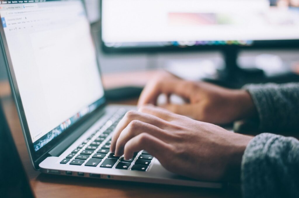 Close-up of hands typing on a laptop keyboard, with a blurred computer monitor in the background. The scene suggests work and leadership in the age of AI, whether working, studying, or writing in an office or home environment.