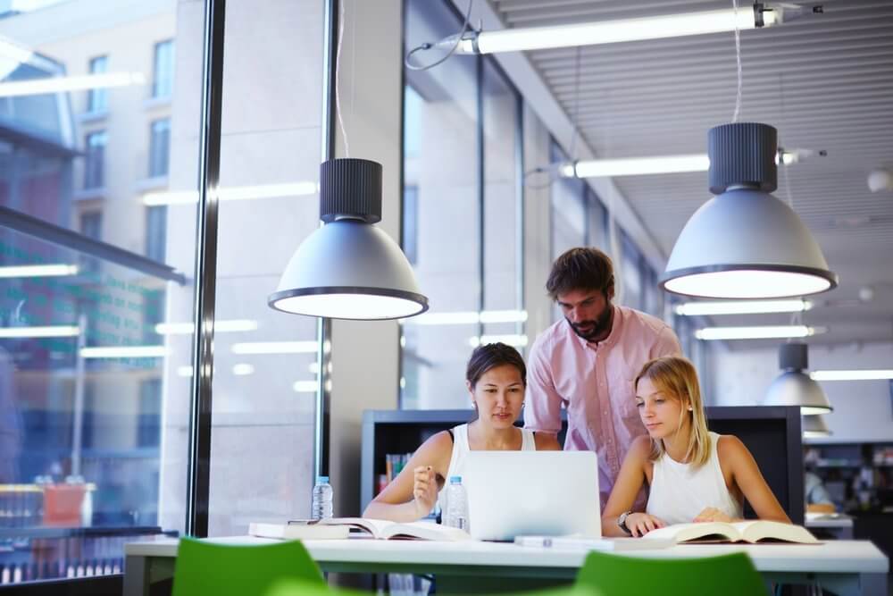 Three people collaborating around a laptop in a modern library with large windows and overhead lights.