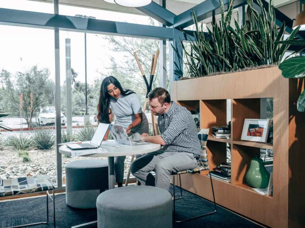 Two hybrid workers collaborate at a round table with laptops in a modern office, surrounded by plants, shelves, and large windows showing trees and parked cars outside.