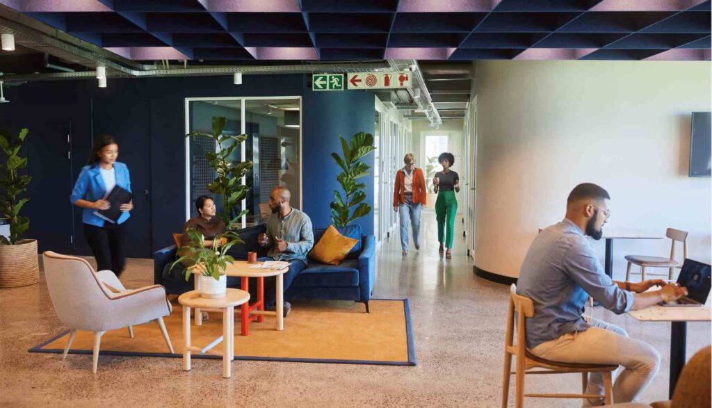 Modern office with people working and talking. Some sit on couches and chairs, others walk in the hallway. Plants decorate the space, reflecting a shift towards sustainability in the future of corporate real estate portfolios. One person types on a laptop at a high table near the foreground.