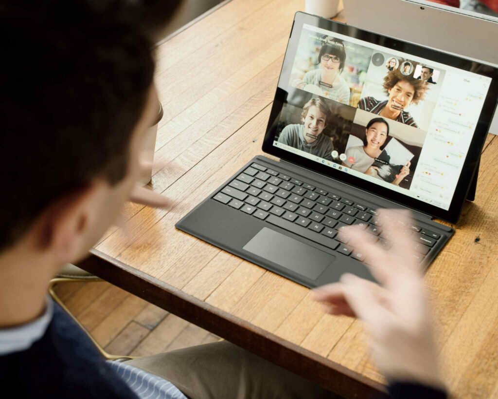 A person gestures in front of a laptop during a video call with five people, all visible on the screen, participating in an online meeting. The setting appears to be a bright workspace with a wooden table—an environment commonly seen in companies with hybrid work models.