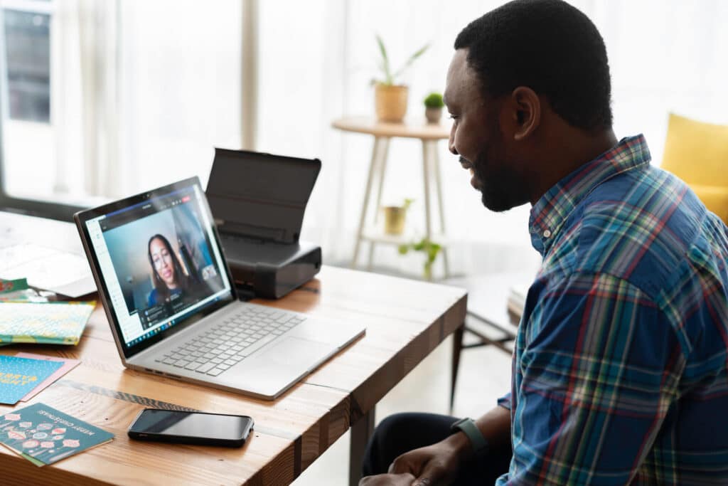Man in plaid shirt facing laptop screen during a video call in a bright room with a plant and papers on the desk.
