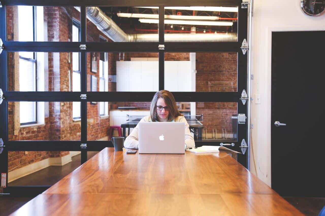 Person working on a laptop at a long wooden table in an industrial-style room with large windows.