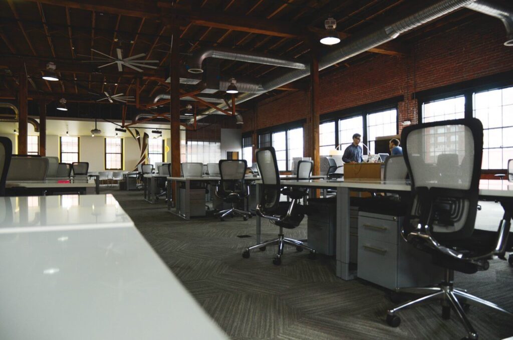 Spacious modern office with rows of desks, chairs, large windows, and wooden beams in an industrial-style space.