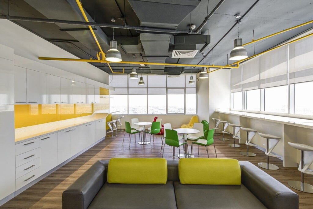 Modern office break room with green chairs, white tables, yellow accents, bar seating, and large windows.