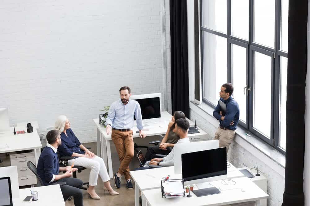 A group of people having a discussion in a modern office with large windows and computer workstations.