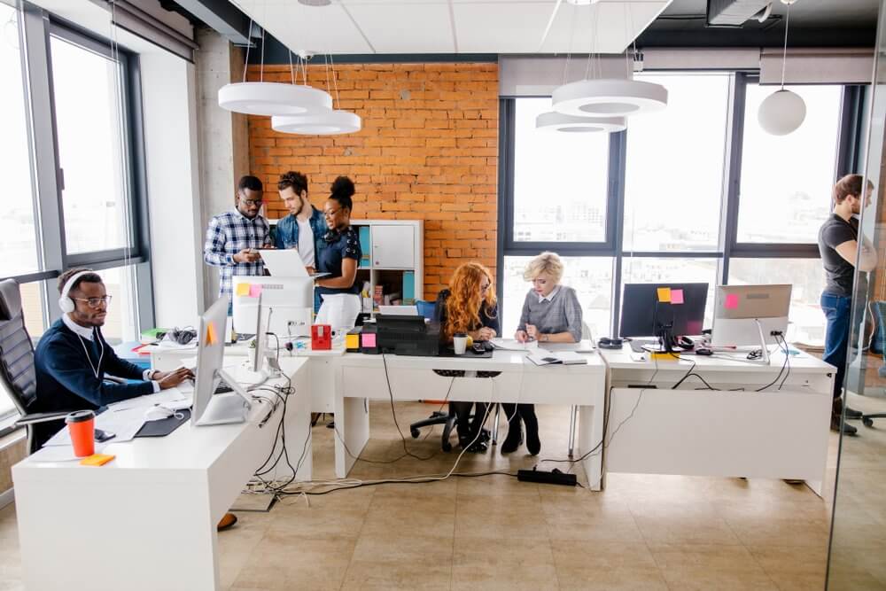 Modern office with diverse team collaborating; some working at desks, others standing and discussing.