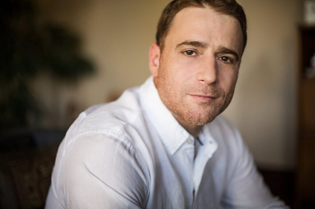 Person with short hair in a white shirt, looking at the camera with a neutral expression indoors.