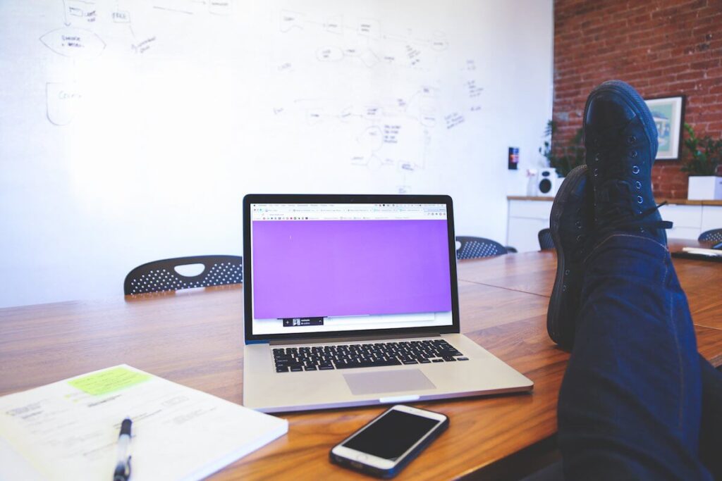 Person relaxing with feet on table, laptop open with purple screen in meeting room, phone and paper nearby.