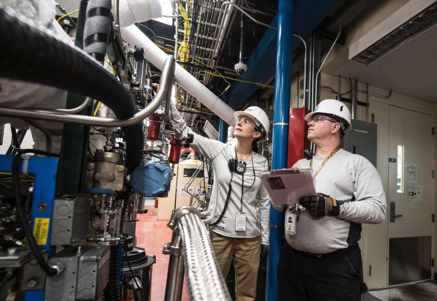 Two engineers wearing hard hats and safety gear examine industrial equipment indoors, focusing on ways to optimize facilities management costs. One points at a component while the other holds a clipboard, surrounded by pipes, wires, and machinery.