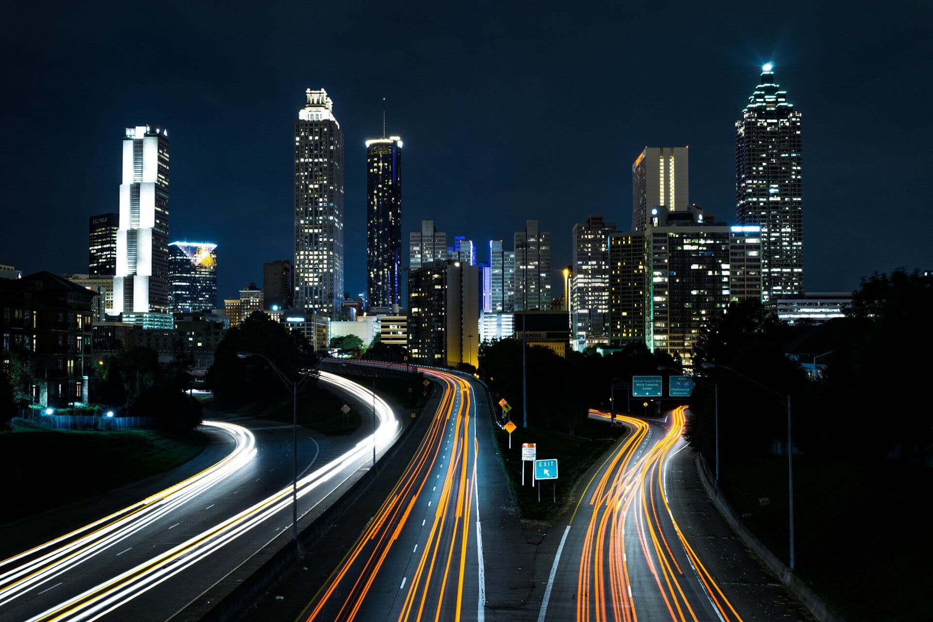 City skyline at night with light trails from traffic on a highway in the foreground.