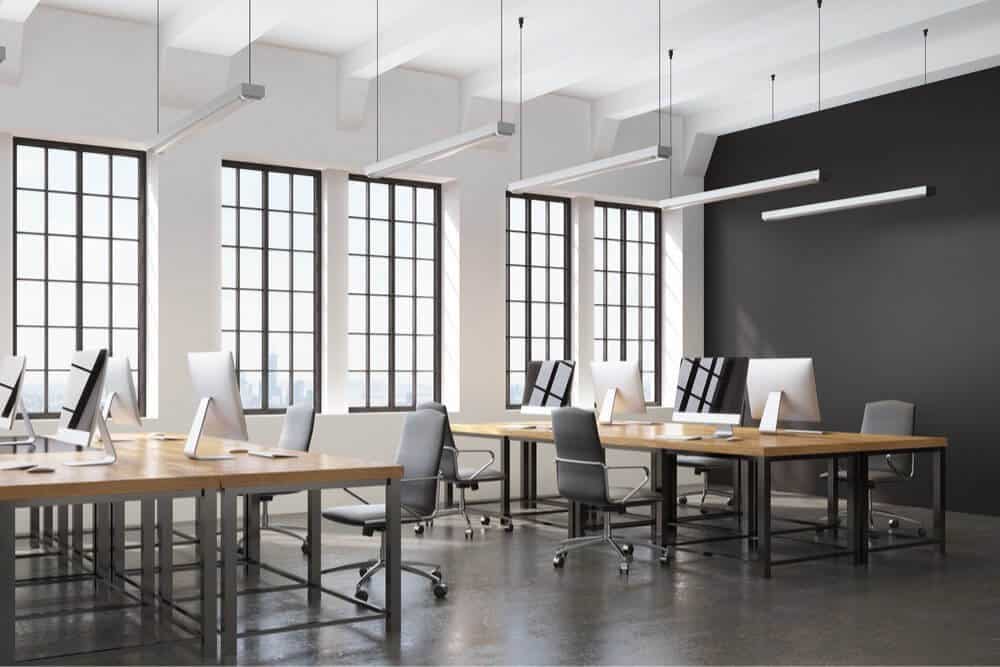 Modern office with large windows, wooden desks, gray chairs, and computers on each desk.