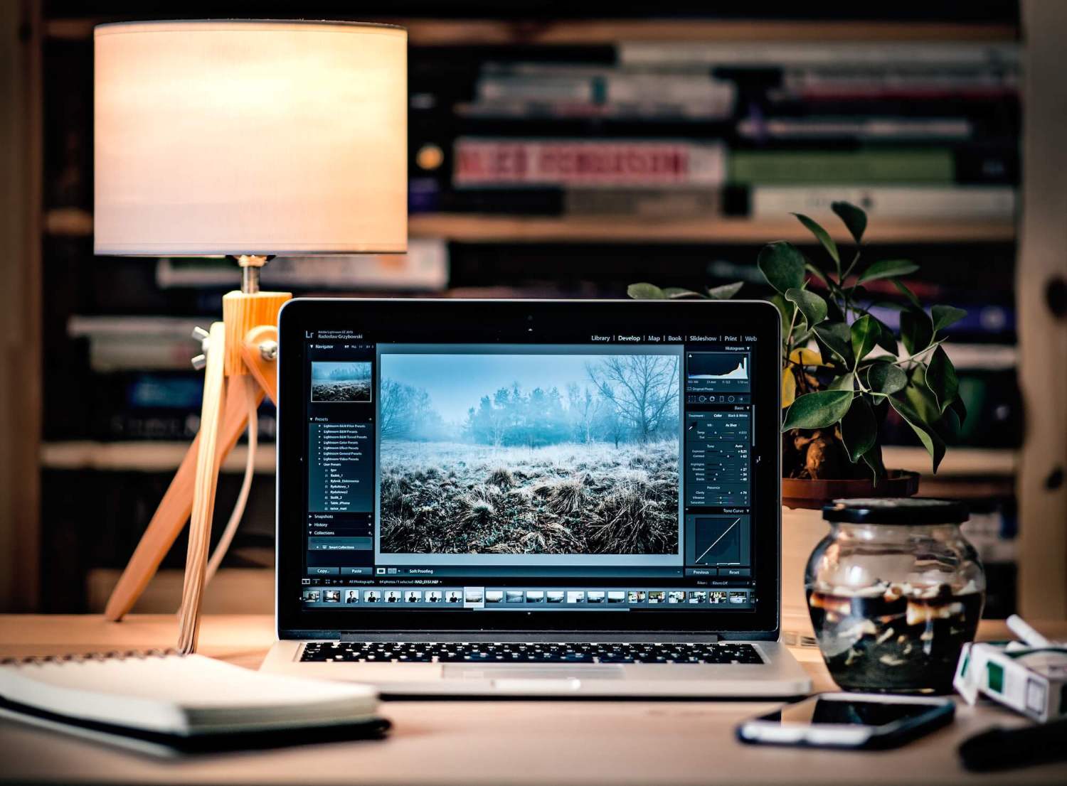 A laptop displaying photo editing software sits on a desk with a lamp, notebook, plant, and glass jar. Books are stacked on shelves in the background, creating a cozy and organized workspace.