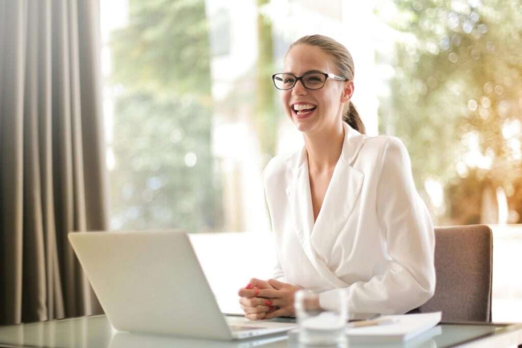 A woman in a white blazer and glasses sits at her desk with a laptop, smiling and laughing as she enjoys the flexibility of hybrid work. Sunlight streams through a window, illuminating the papers and a glass of water on her desk.