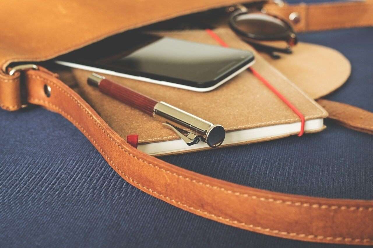 A brown leather bag with a strap lies open, revealing a notebook, a pen, a smartphone, and sunglasses inside, all resting on a dark fabric surface.