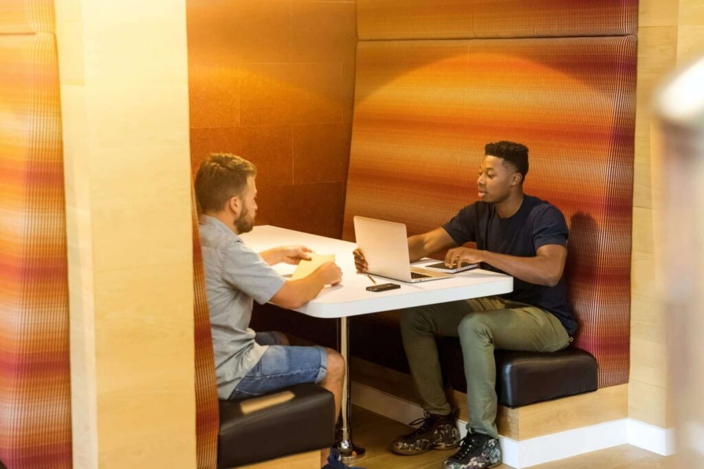 Two men sit across from each other at a table in a booth, one using a laptop and the other taking notes, appearing to have a discussion in a modern, warmly lit workspace designed for activity-based working.