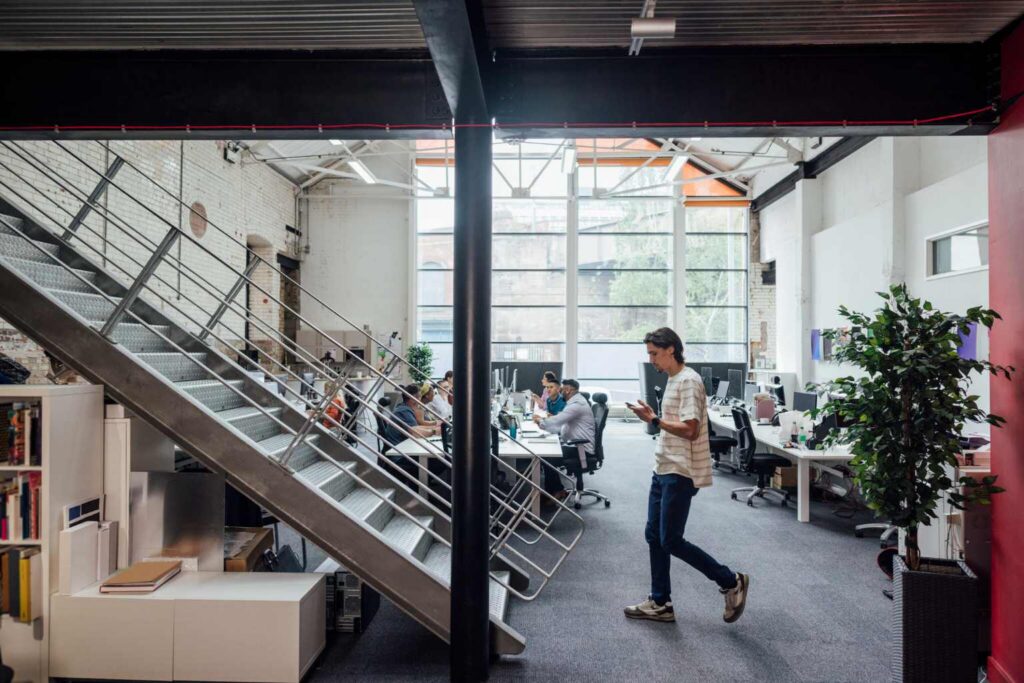 A modern office with people working at desks. A person walks near a metal staircase, and large windows are in the background.
