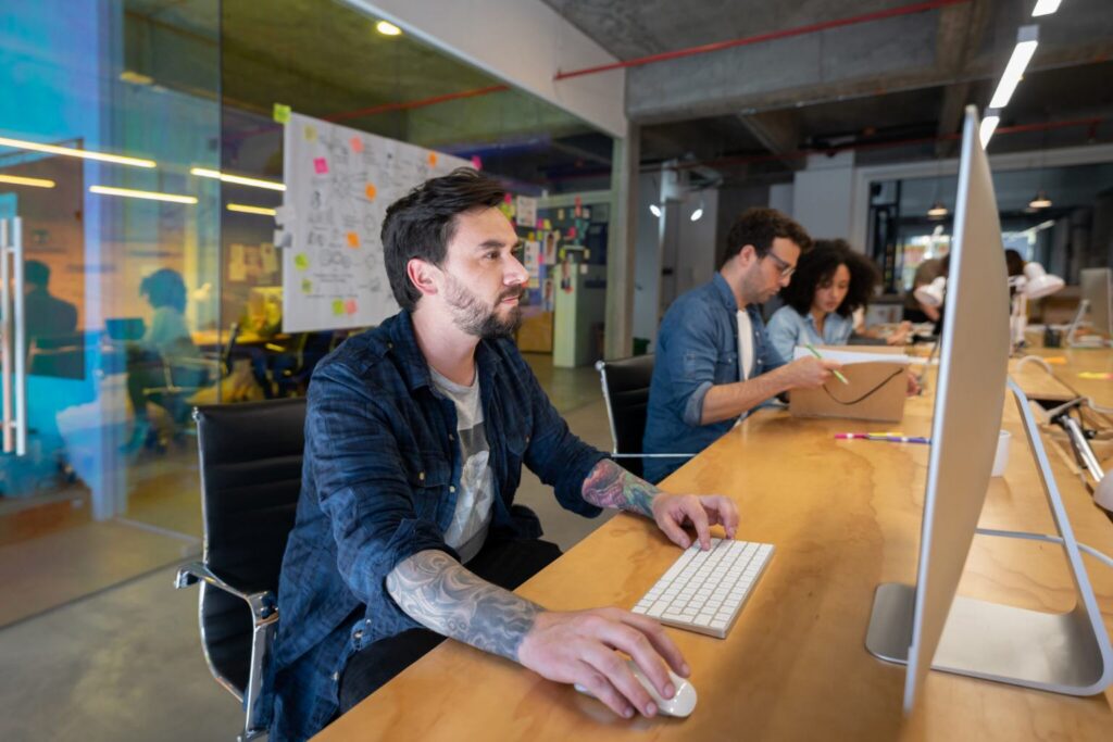 Three people working at computers in a modern office with glass walls and various notes on a board behind them.