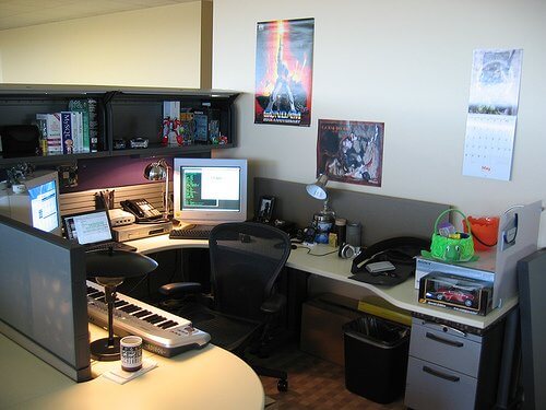 A cluttered office cubicle with a keyboard, computer monitors, various decorations, and a calendar on the wall.