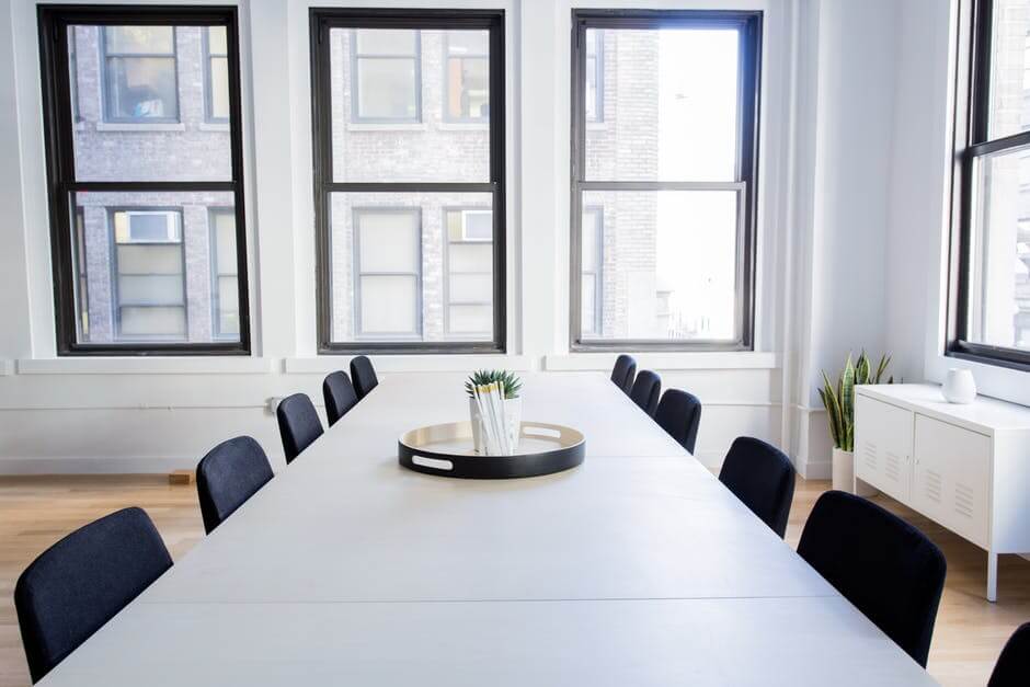 A modern conference room with a long table, black chairs, large windows, a tray with pens, and a potted plant.