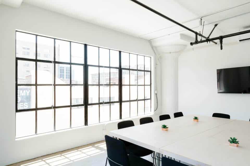 Bright meeting room with large windows, a long table, black chairs, and a wall-mounted TV.
