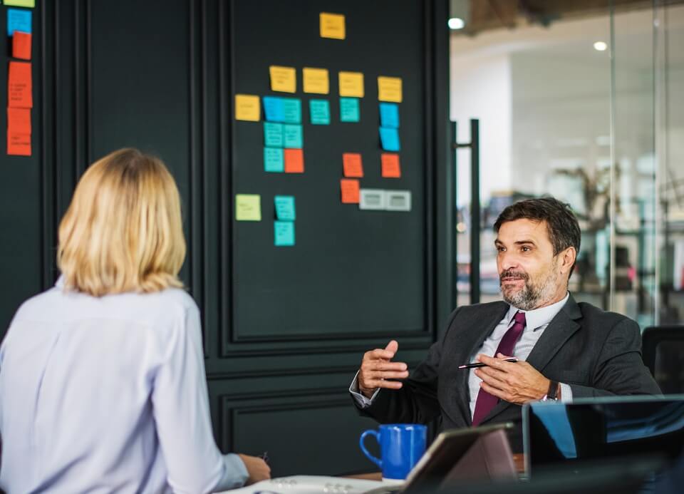 Two people having a discussion in an office, with sticky notes on a blackboard in the background.