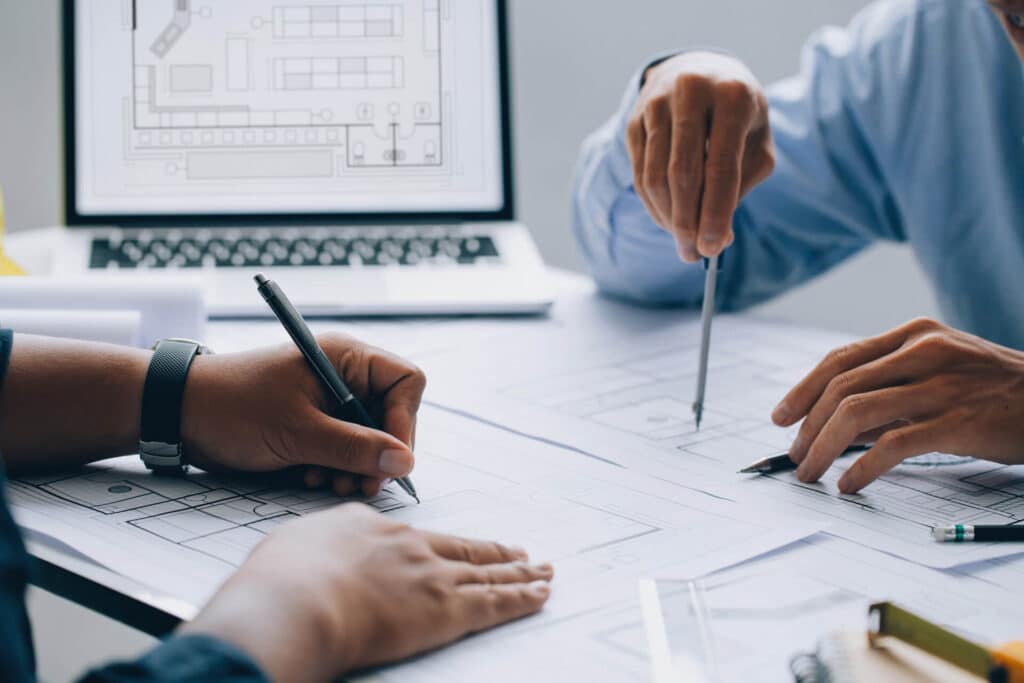 Two people work on architectural blueprints with a laptop displaying designs in the background.