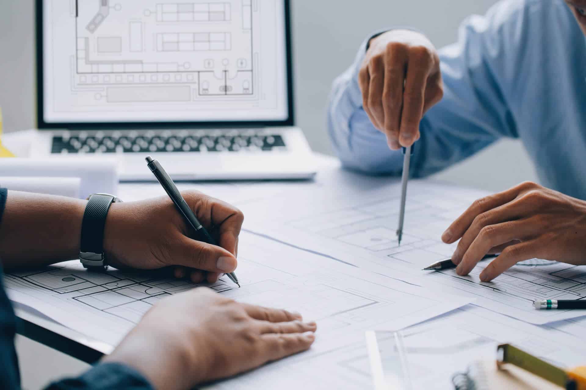 Two people work on architectural blueprints with a laptop displaying designs in the background.