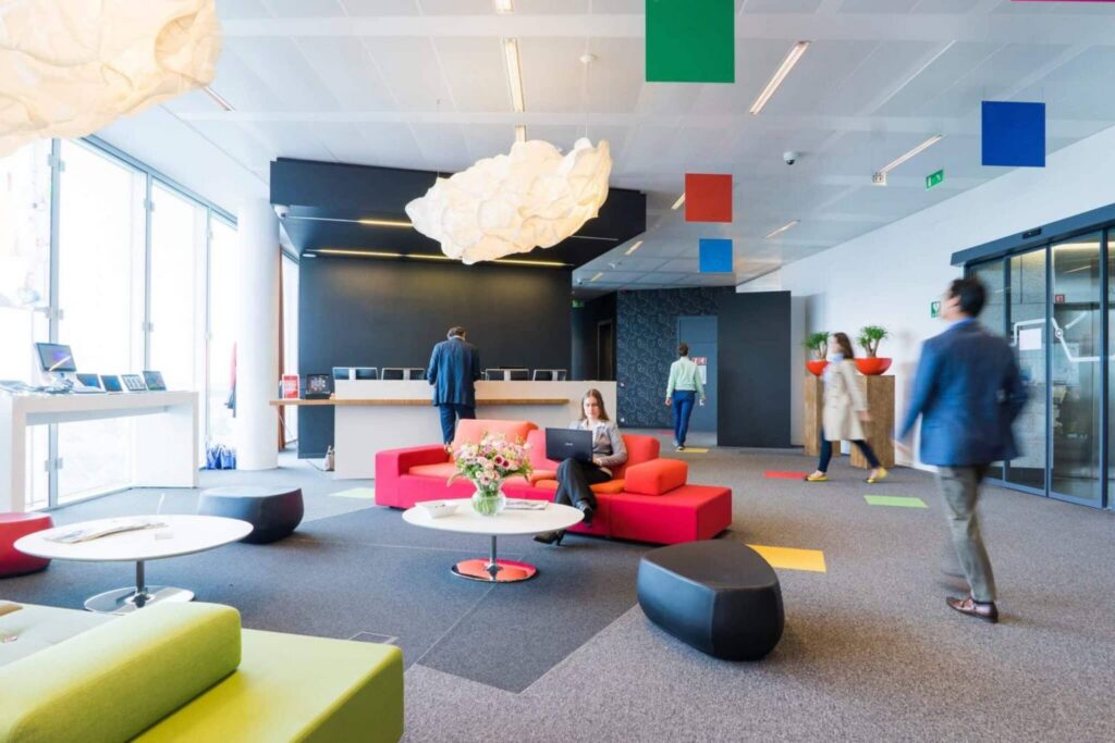 Modern office lobby with colorful furniture, geometric ceiling decorations, and people walking or sitting—showcasing an activity-based working environment. Large windows let in natural light, and a reception desk is visible in the background.