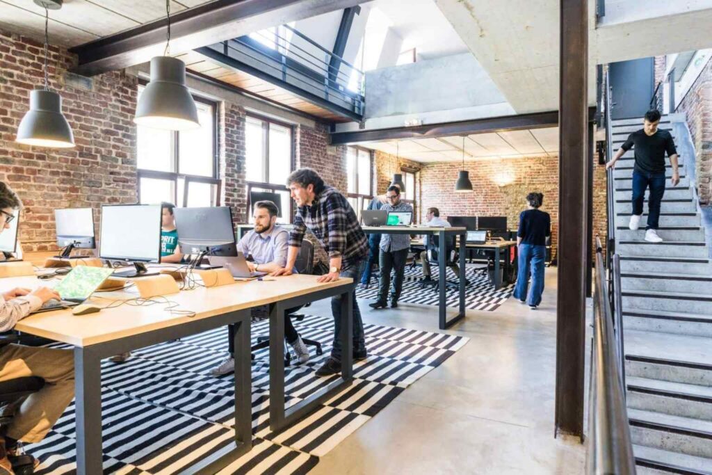 Modern open-plan office with people working at desks, collaborating, and using computers—showcasing agile work examples in action. Exposed brick walls, large windows, black-and-white rugs, and a person walking downstairs are visible in this bright, spacious environment.