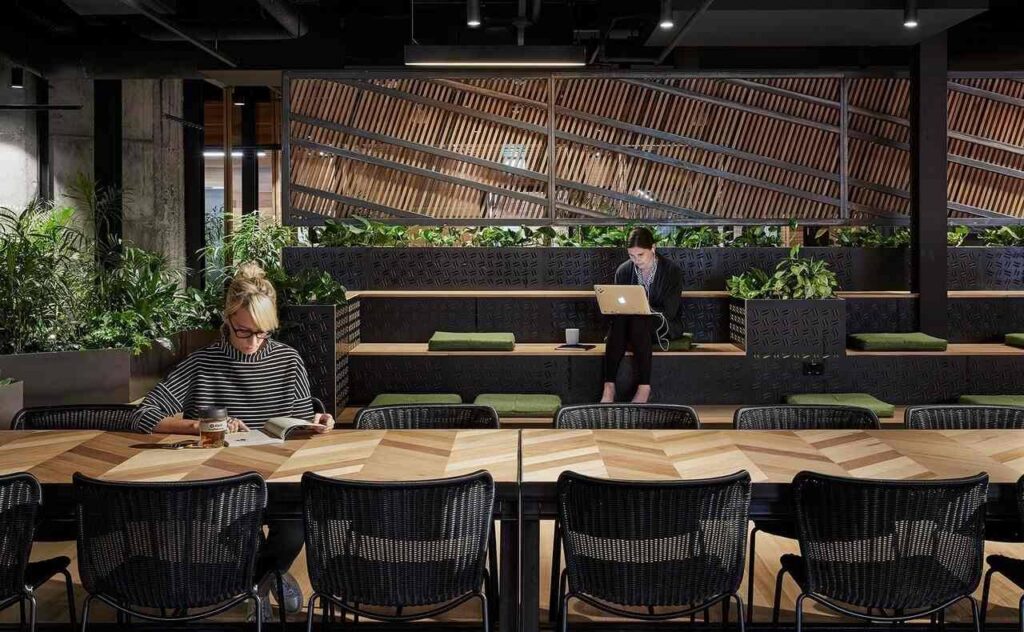Modern workspace with wood tables, black chairs, tiered seating with green cushions, lush plants, and two people—one reading at a table, another using a laptop on the steps. Geometric wood paneling decorates the wall.