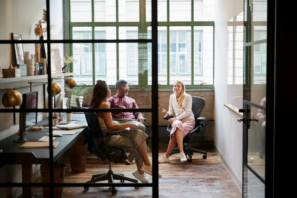 Three people having a meeting in a modern office with large windows and natural light.