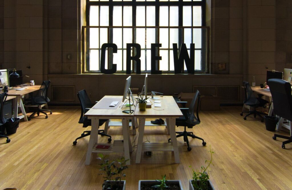 Open office space with wooden desks, computers, and large "CREW" letters by a window. Plants in the foreground.
