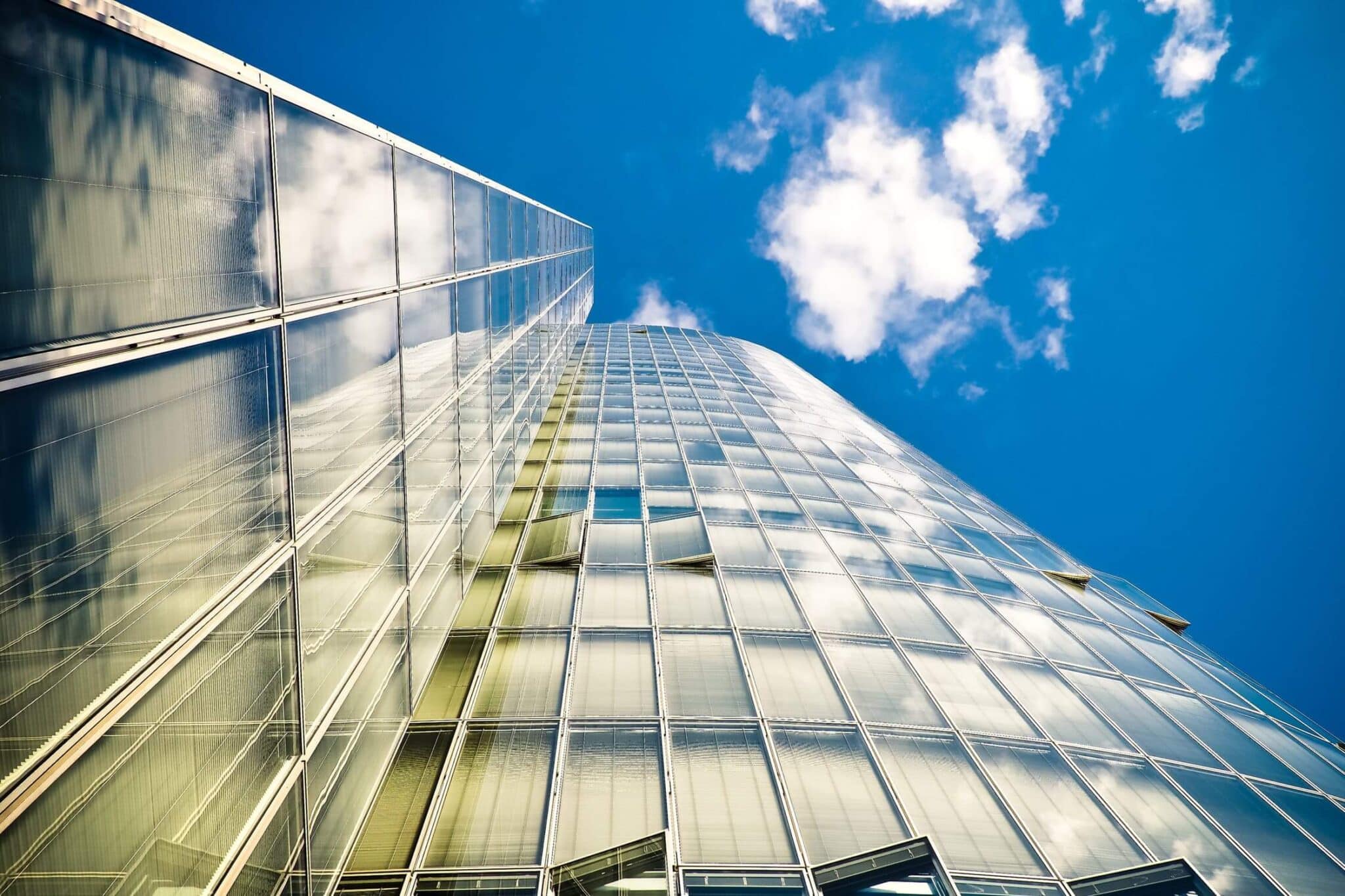 Upward view of a glass skyscraper reflecting clouds and blue sky.