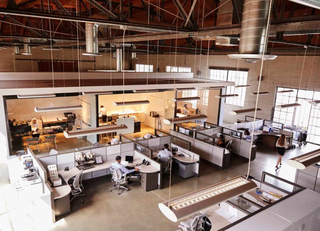 Open-plan office with employees at cubicles, high ceiling, exposed beams, and modern lighting.