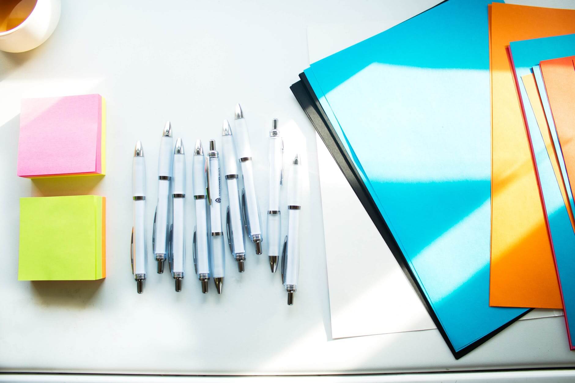 Colorful sticky notes, pens, and folders on a white desk, with sunlight casting shadows.