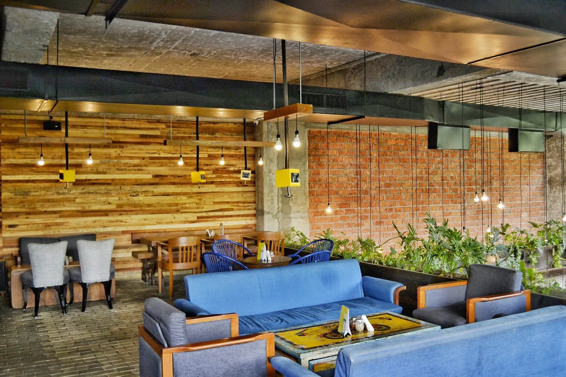 Modern cafe interior with blue sofas, wooden tables, hanging lights, and exposed brick walls.