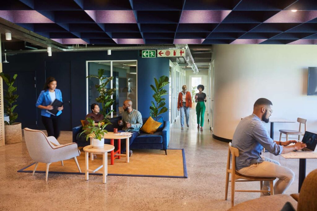 Modern office with people working, walking, and sitting on sofas. Plants and signage are visible in the spacious area.