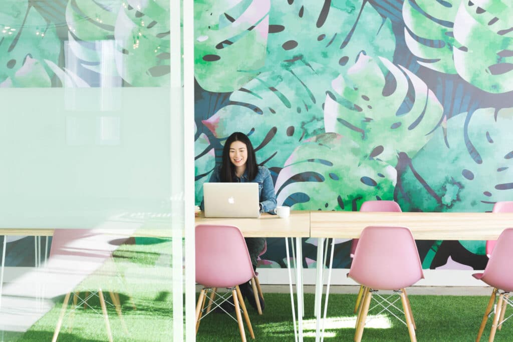 Person working on a laptop at a table with pink chairs, set against a leafy mural background.