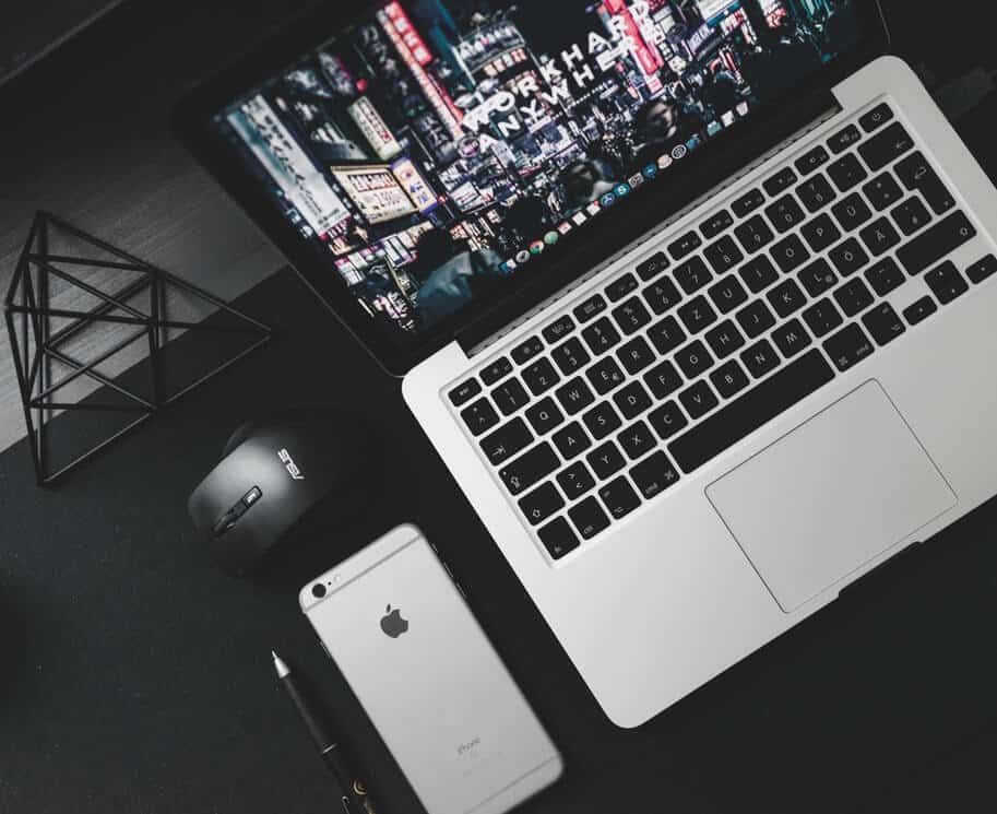 A laptop displaying a cityscape, with a smartphone, stylus, and computer mouse on a dark desk.