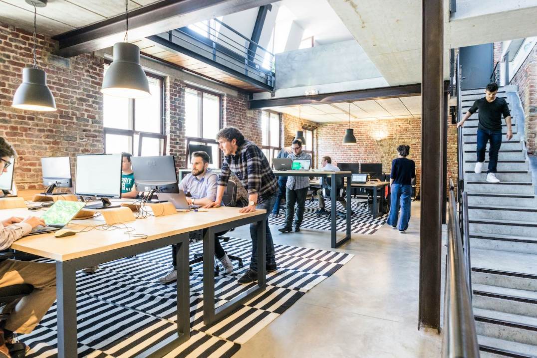 Modern open-plan office with people working at desks, collaborating, and using computers—showcasing agile work examples in action. Exposed brick walls, large windows, black-and-white rugs, and a person walking downstairs are visible in this bright, spacious environment.