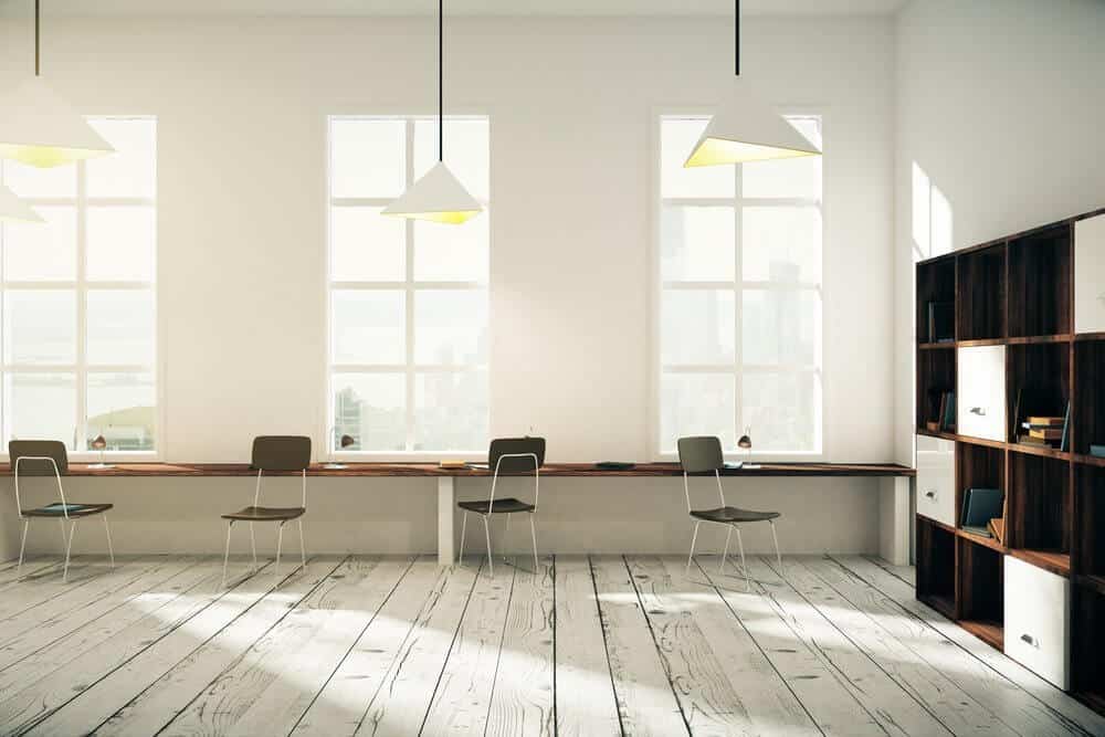 Bright minimal office space with wooden desks, chairs, large windows, pendant lights, and a bookcase.
