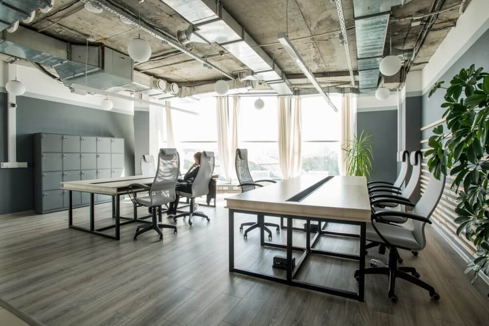 Modern office with wooden tables, black chairs, overhead lighting, large windows with curtains, and plants.