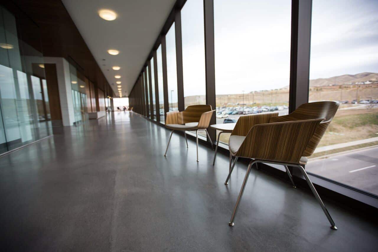 Modern corridor with large windows, two wooden chairs, and a view of a parking lot and hills outside.