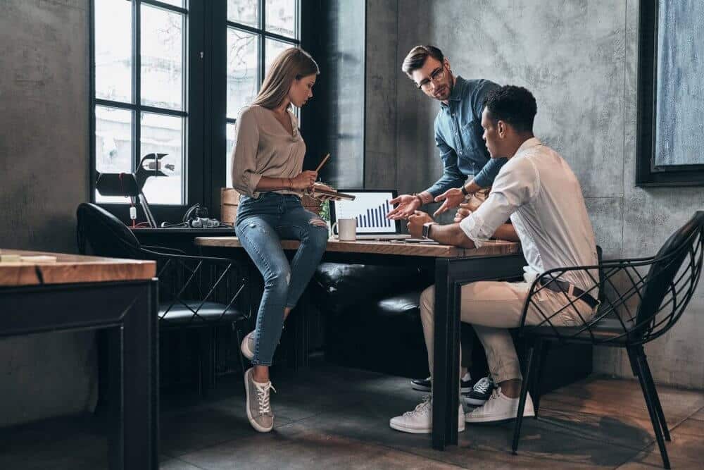 Three people in a modern office discussing a bar graph on a laptop screen at a wooden table.