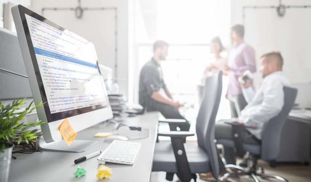 Close-up of a computer monitor displaying code, with four people collaborating in the background of an office setting.