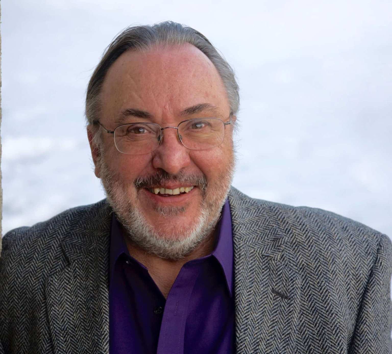 Smiling man with glasses, wearing a purple shirt and gray herringbone jacket, against a light background.
