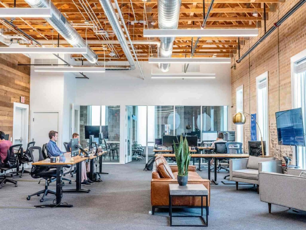 Open modern office with exposed ceilings, desks with computers, several people working, a brown leather sofa, potted plant, and glass-walled meeting rooms in the background. Reflecting the principles of OfficeSpace's Maturity Model, the environment is thoughtfully designed to foster collaboration and productivity. Natural light enters through tall windows.