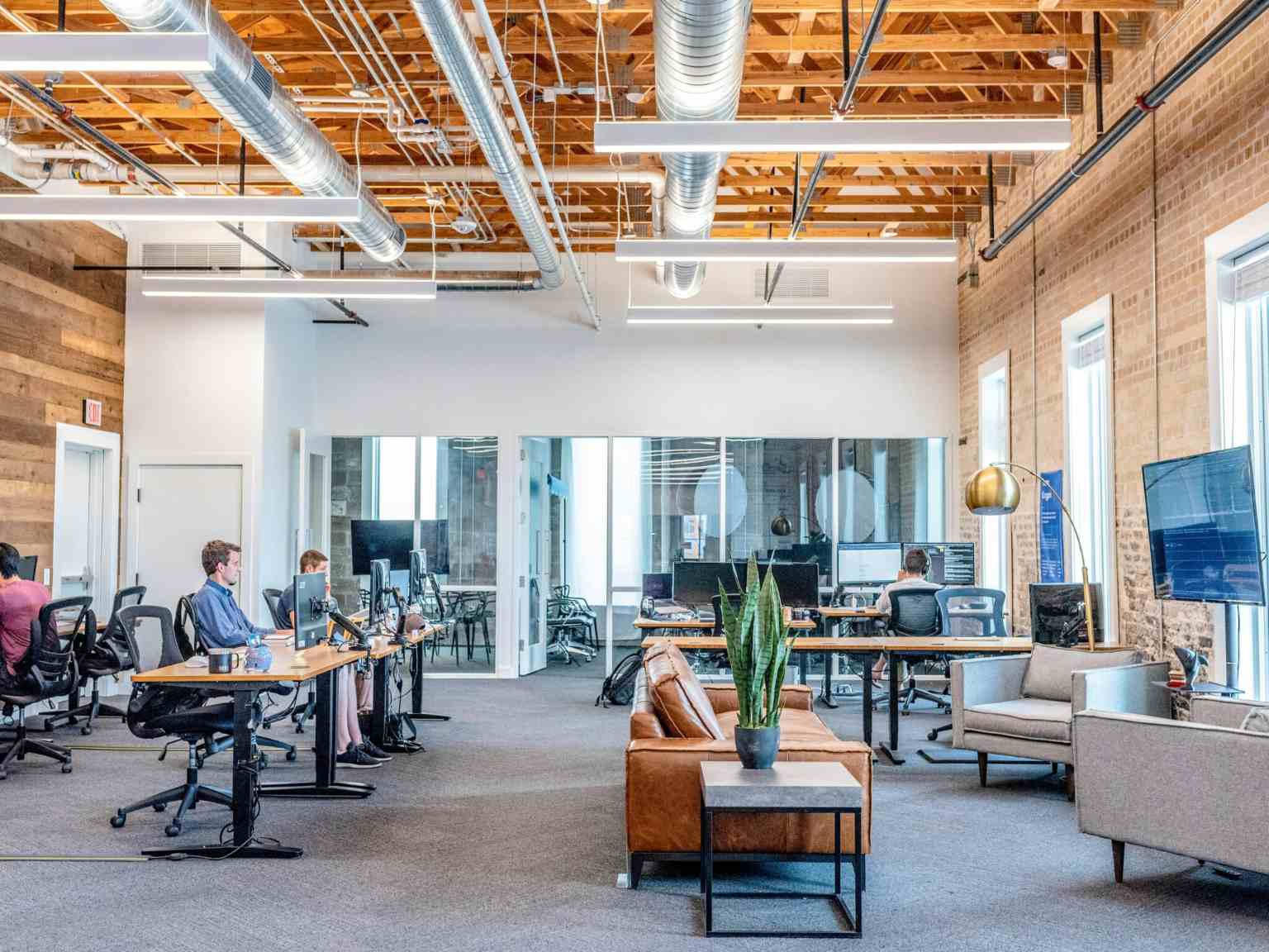 Modern open office space with exposed ceilings, glass-walled meeting rooms, desks with computers, people working, a brown leather sofa, green plant, and large windows letting in natural light.