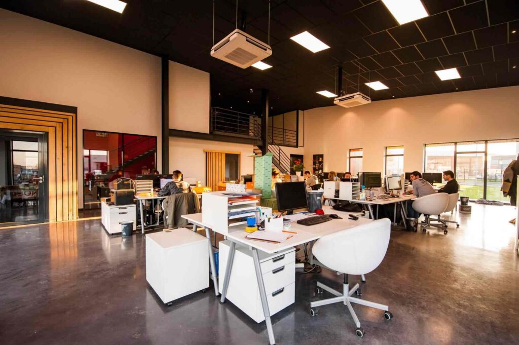 Modern open office space with desks, computers, and people working. Large windows let in natural light. Some staff are seated at desks while others move around. The room has a high ceiling and a mezzanine level.